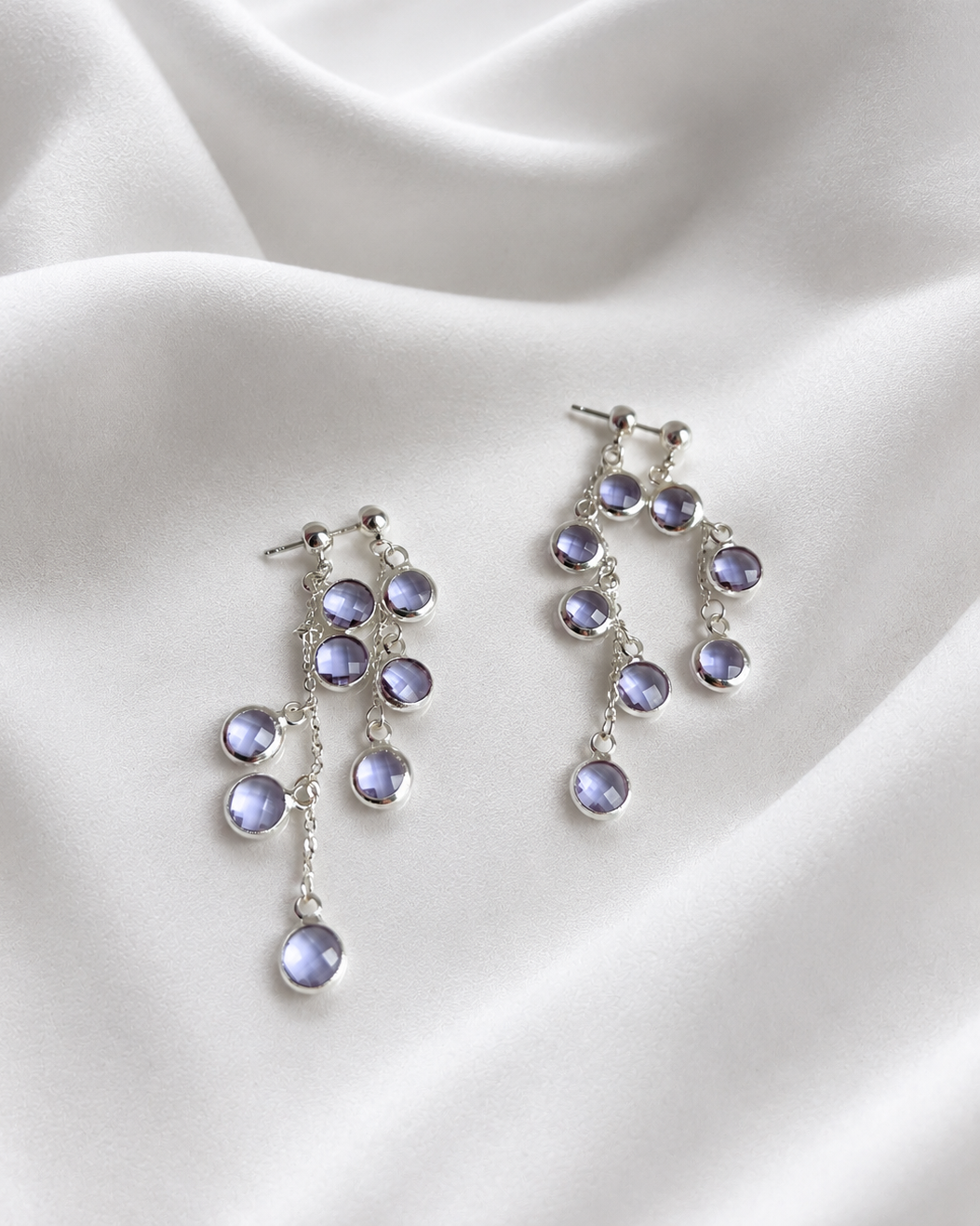 A pair of silver drop earrings with multiple small oval purple gemstones, displayed on draped white satin fabric.