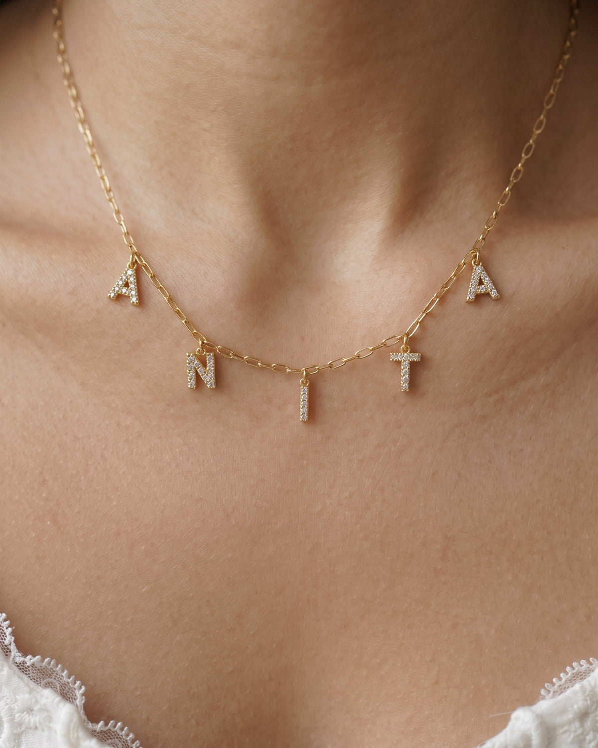 A close-up of a person's neck and upper chest wearing a delicate gold chain necklace with letter charms spelling 'ANTA'. The letters are adorned with small clear stones, and the necklace is shown against warm skin tones with a white lace garment visible at the bottom.