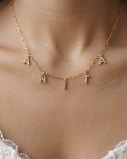A close-up of a person's neck and upper chest wearing a delicate gold chain necklace with letter charms spelling 'ANTA'. The letters are adorned with small clear stones, and the necklace is shown against warm skin tones with a white lace garment visible at the bottom.