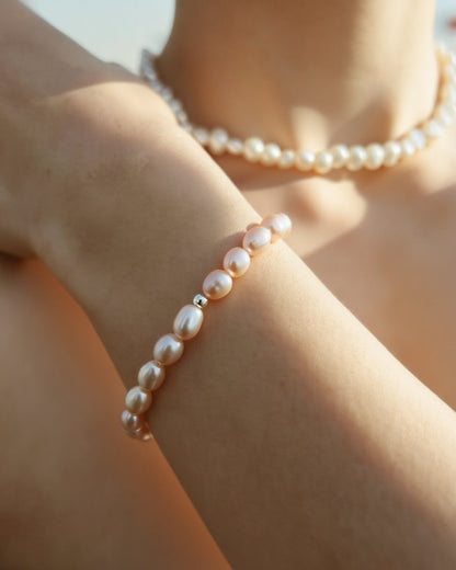 A close-up of a person's wrist wearing a delicate bracelet made of small white pearls separated by tiny metallic spacer beads, with a matching pearl necklace visible in the background.