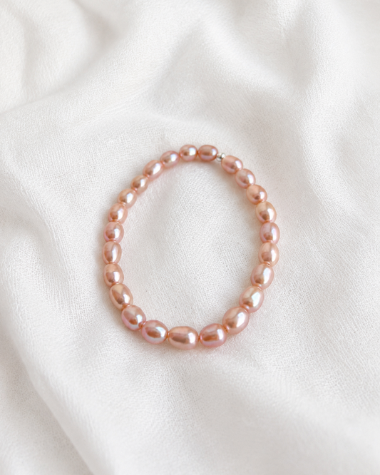 A single strand bracelet made of small, irregular, glossy pink freshwater pearls, arranged in a loose circle on a soft, draped white fabric background.