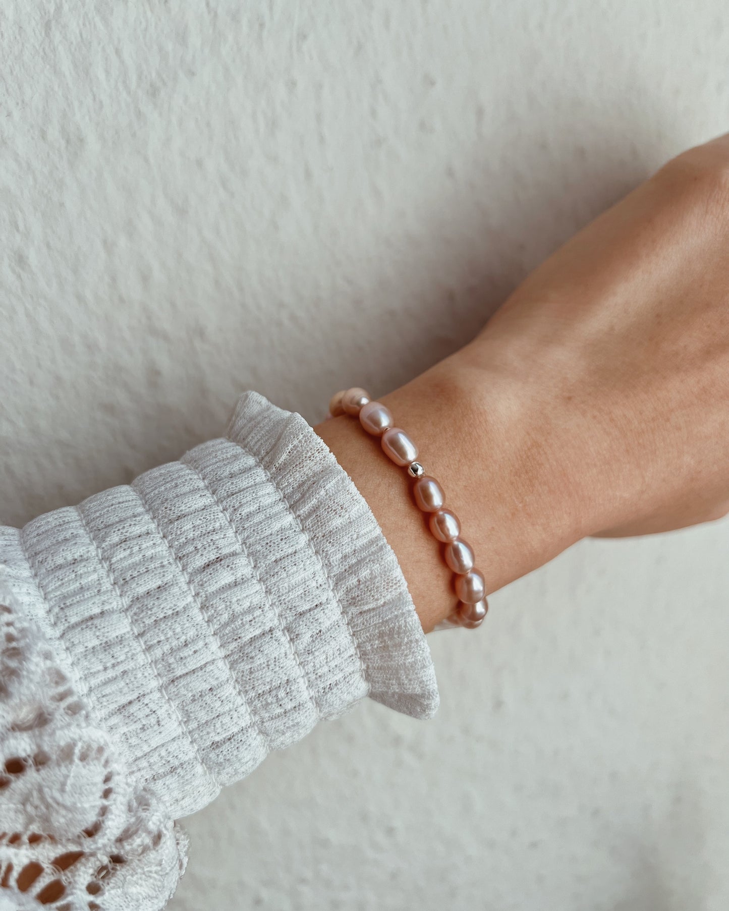 A close-up of a person's wrist wearing a delicate beaded bracelet made of small, glossy dusty pink pearls, styled with a white ribbed knit sleeve.