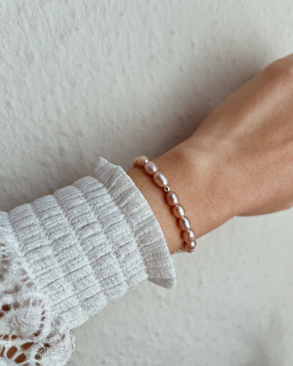 A close-up of a person's wrist wearing a delicate beaded bracelet made of small, glossy dusty pink pearls, styled with a white ribbed knit sleeve.