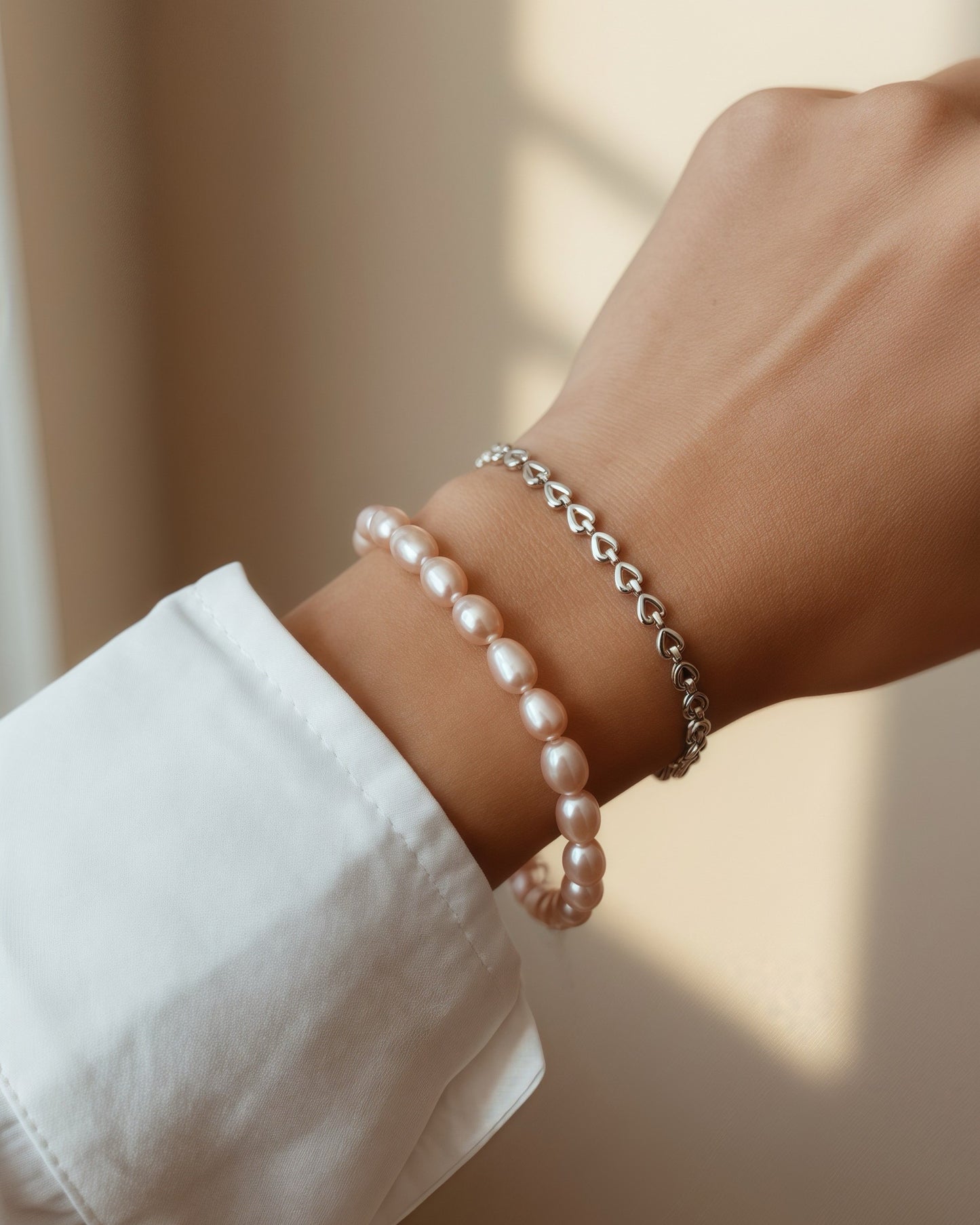A close-up of a person's wrist wearing two delicate bracelets: a strand of light pink pearl-like beads and a thin silver chain bracelet with heart-shaped links, shown against a softly lit beige background.