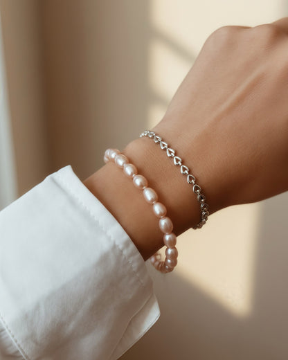 A close-up of a person's wrist wearing two delicate bracelets: a strand of light pink pearl-like beads and a thin silver chain bracelet with heart-shaped links, shown against a softly lit beige background.