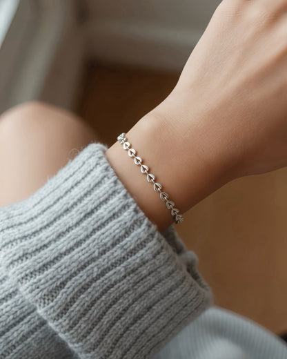 A delicate silver chain bracelet with small leaf-like links worn on a person's wrist, paired with a gray ribbed knit sweater sleeve.