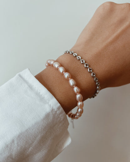 A close-up of a person's wrist wearing two delicate bracelets: one strand of lustrous light pink pearls and one silver chain bracelet with small heart-shaped links, against a soft light gray background.