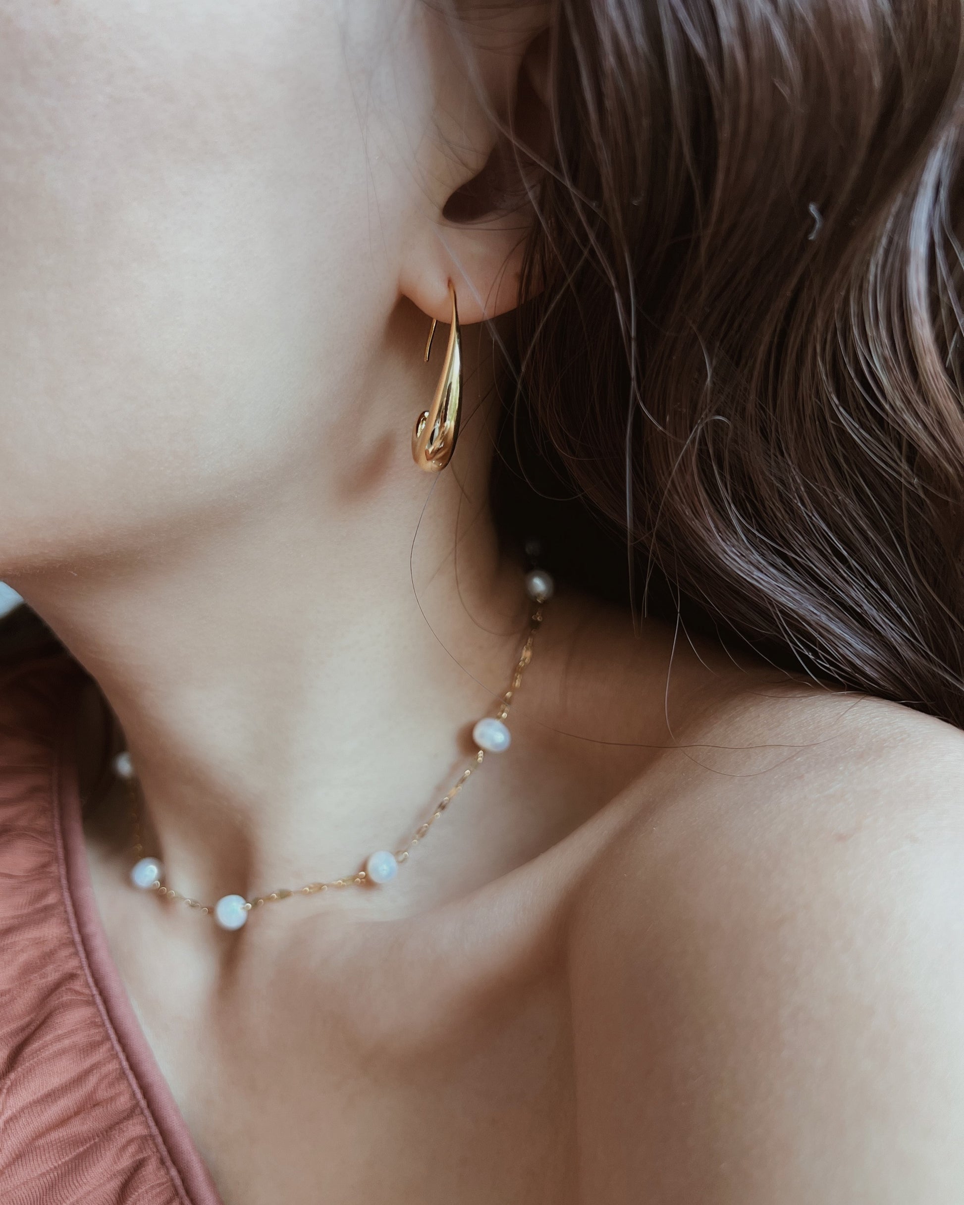 Close-up of a woman wearing a gold huggie hoop earring and a delicate gold necklace with white pearl-like beads.