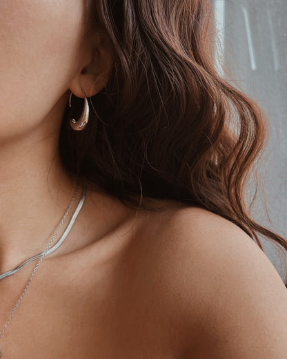A close-up lifestyle shot of a woman wearing a polished silver crescent moon hoop earring, layered with a delicate silver chain necklace. She has wavy brown hair and is shown from the side against a soft gray background.