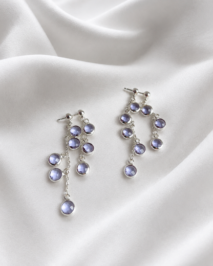 A pair of silver drop earrings with multiple small oval purple gemstones, displayed on draped white satin fabric.